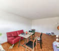 Red leather couch, wooden table, 2 chairs, books, plant, floor lamp, white walls, wooden floor