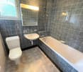The image shows a bathroom with gray and black tile walls, a white toilet, a white sink, and a white bathtub. The bathroom has a window and lighting fixtures.