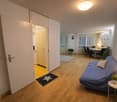modern living room, hardwood floors, open-plan layout, natural light, simple decor, comfortable seating, desk, potted plants