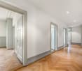 The image shows a spacious hallway with white walls, hardwood herringbone flooring, and several doorways leading to other rooms. The hallway has a clean, modern aesthetic with recessed lighting and neutral color tones.