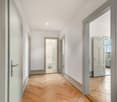 Bright and spacious hallway with hardwood herringbone flooring, white walls, and multiple doors leading to other rooms. The hallway appears to be part of a larger apartment or home.