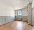 Spacious room with high ceilings, hardwood herringbone flooring, and white walls with wainscoting paneling. Large window overlooking a view of neighboring buildings.