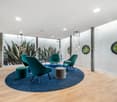 Spacious office lounge with large wall mural of tropical plants, teal armchairs and ottomans, round blue rug, wooden flooring, and circular wall mirrors providing a view of the outdoors.