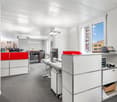 modern office space with white walls, gray carpet, white desks, red and white cabinets, computer monitors, printers, and office equipment