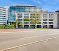 Multi-story commercial building with glass facade, surrounded by trees and located on a busy street with parking spaces