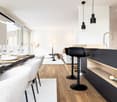 Dining area with a large table, chairs, wooden floor, modern white wall, a white rug, and black stools with a modern faucet.