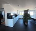 Open kitchen with white cabinets, black countertop, modern fixtures, wooden dining table, chairs, plant, large windows