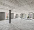 Large open-plan space with concrete ceiling and floor, glass sliding doors leading to a balcony or terrace, and exposed concrete columns.