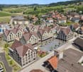 Aerial view of a picturesque town with traditional European-style buildings, red-tiled roofs, and a central plaza. The town is surrounded by lush green hills and countryside. There are several multi-story apartment buildings and a parking area visible in