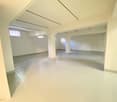 Large open space with high ceilings, white walls, and concrete flooring. The room appears to be an unfinished or renovated space, with exposed beams and windows providing natural light.