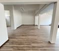The image shows a large, open office space with a high ceiling and white walls. The floor is covered in a wood-like laminate flooring. The space appears to be empty and ready for use, with track lighting installed overhead.