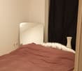 Bed with brown sheet, wooden floor, wall mounted mirror, white wall, curtain and a white vase