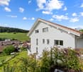 Detached 2-storey house, white, with brown roof, 4 windows, located in a lush environment with trees and grass