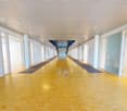 Long, well-lit hallway with white walls, a tiled yellow floor, and multiple doors on both sides. The hallway has a high ceiling with recessed lighting and appears to be part of a larger commercial or institutional building.
