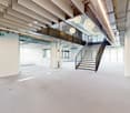 Large open-plan office space with high ceilings, exposed beams, and modern lighting fixtures. The space features a central staircase leading to a mezzanine level, and there are glass walls separating different areas. The floor is covered in a light-colore