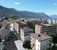 Aerial view of a town with several buildings, mountains in the background, solar panels on some buildings, cars parked on the street, trees and greenery