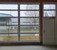 The image shows a commercial building with large windows overlooking a parking area and some trees outside. The building has a gray exterior and the windows have a modern, industrial-style design. There appears to be a door on the right side of the buildi