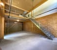 The image shows a spacious garage or storage area with a wooden ceiling, concrete floor, and a metal staircase leading to an upper level. The space appears to be unfinished, with exposed wooden beams and OSB board walls.