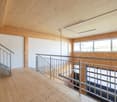 The image shows a large open space with a wooden ceiling and walls. There is a metal railing along the upper level, indicating a second floor or mezzanine. The floor is made of wood planks. The space has large windows that provide natural light. Overall,