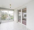 Spacious balcony with glass sliding doors, tile flooring, white walls, balcony railing, trees and building views outside