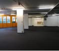 Empty room with ceiling lights, white columns, windows, and a door. Floors are carpeted. Another room is visible in the background.