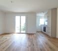 Empty room with sliding glass door to balcony, white walls, wooden floor, kitchen with oven and sink