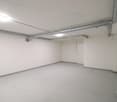 Empty garage room, white walls, ceiling, gray concrete floor, white ceiling lights, pipe installation on ceiling and walls, and a white door.