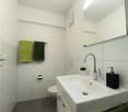 white walls, white tiles, white sink, chrome faucet, white toilet, green towels, small plant, mirror, soap dispenser