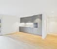empty kitchen with built-in appliances, light wooden flooring, sliding glass door, white walls, gray cabinetry, kitchen island with sink