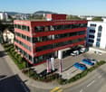 Red office building, multiple floors, multiple windows, parking lot with several cars, street light, greenery