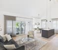 modern living room with large windows, glass doors, wooden floor, gray couch, glass coffee table, pendant lights, kitchen island, dining table, and chairs