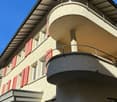 2-story building with a curved facade, beige exterior walls, red shutters on the windows, and a balcony on the upper level.