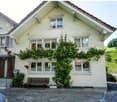 A white, two-story house with a brown roof and white windows covered by climbing plants. There is a small garden with a wooden bench and plants to the right of the house. The front has brick-paved driveway and a parked car. The house has steps leading to