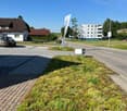 The image shows a residential neighborhood with a mix of single-story houses and multi-story apartment buildings. There is a paved walkway and a grassy area with some vegetation. A street lamp and a flag pole with a banner are visible. The sky is clear an