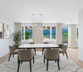 Spacious dining room with large windows offering a scenic view of the surrounding landscape. The room features a marble dining table surrounded by plush gray chairs, and modern pendant lighting fixtures. The walls are adorned with framed artwork, and a po