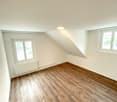 Empty room with wooden floors, two windows, sloped ceiling with integrated lighting, heating radiators under windows.