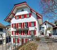Large multi-story house with red shutters and balconies, fenced entrance, statue, gravel path, and small garden