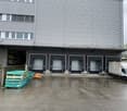 The image shows a large industrial building with multiple loading dock doors. There are stacks of pallets and a delivery van parked outside, indicating this is likely a warehouse or distribution center.