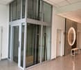 The image shows an interior office space with a glass elevator or lift in the center. The walls are white, and the floor is made of granite or a similar material. There is a large circular light fixture on the wall, and some small benches or tables near t