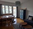 A room with wooden flooring and large windows, dining table with chairs, wooden cabinet, sofa with pillows, and wooden tables.