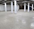 Large open space with concrete flooring, white support columns, and overhead lighting. The space appears to be empty and unfinished, likely a warehouse or industrial facility.