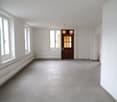 Large open space with white walls, hardwood floors, and a wooden door. The room has large windows providing ample natural light.
