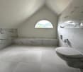 Spacious bathroom with marble-like tiles covering the walls and floor, a large arched window providing natural light, a modern oval-shaped sink, and a toilet.