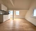 The image shows a studio apartment with an open floor plan. The room has hardwood floors, white walls, and large windows that provide natural light. The kitchen area is visible, with white cabinets and appliances. The overall space appears bright, clean,