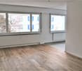 This image shows an empty room with large windows overlooking a snowy outdoor scene. The room has a hardwood floor and white walls. There is a radiator or heating unit visible along the wall.