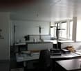 The image shows an office space with a desk, computer equipment, and various office supplies. The room has large windows providing natural light, and the walls are painted a light color. The overall layout and furnishings suggest a professional work envir