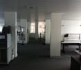 Large open office space with white walls, black carpeting, and various office equipment such as printers, computers, and a refrigerator. There appears to be a private washing machine and dishwasher visible in the room.