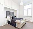 white bedroom with grey headboard, 3 drawers, 2 nightstands, bench at foot of bed, radiator, window, grey carpet