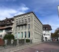 4 story residential building with multiple balconies, brown roof, large windows, flag on the building