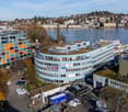 An aerial view of an office building, featuring several floors with multiple windows, surrounded by trees and a busy street with multiple cars parked in front. Other buildings, including another office building, are visible in the distance, along with a l